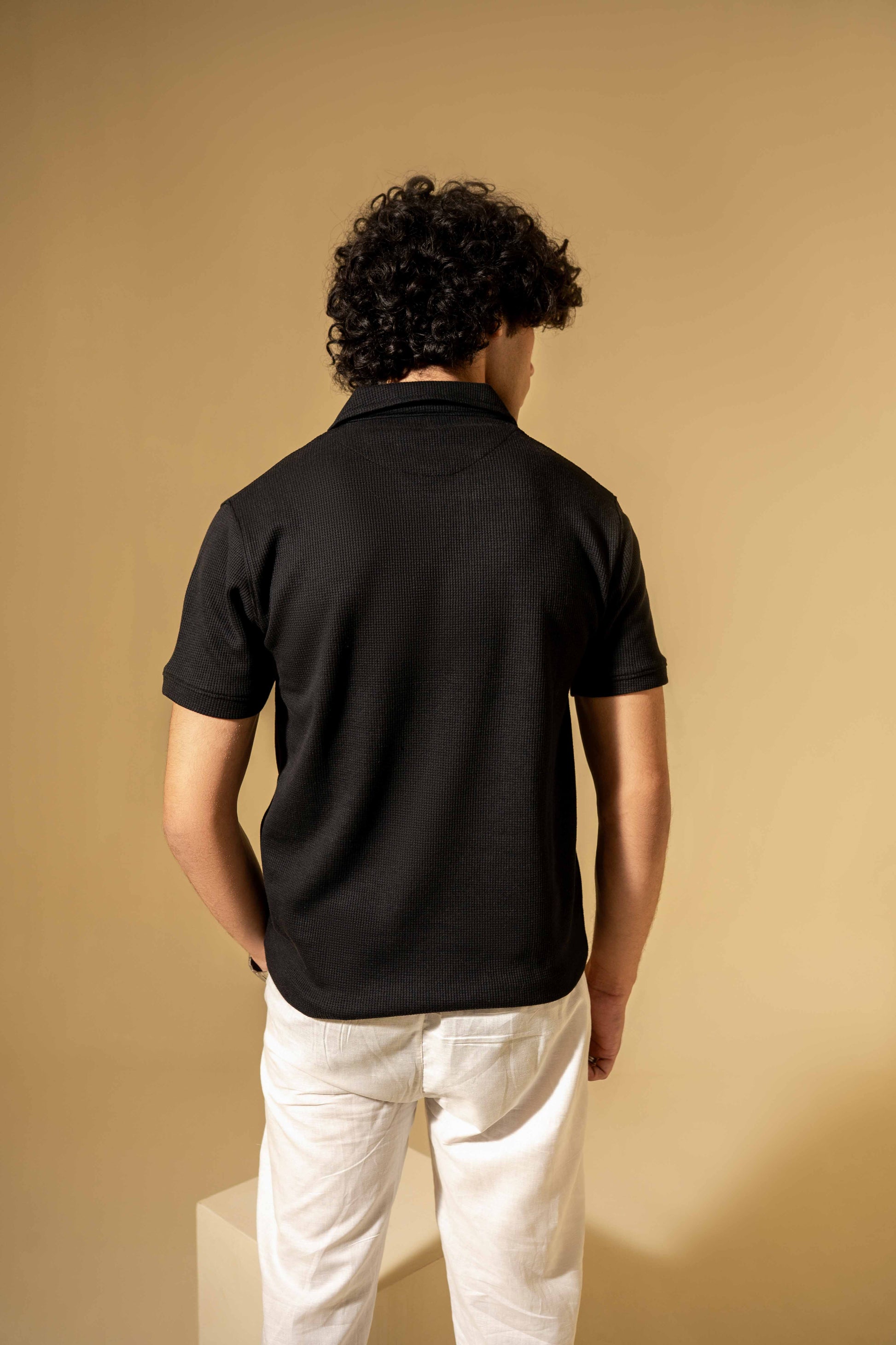 Person wearing a black polo shirt and white pants against a beige background