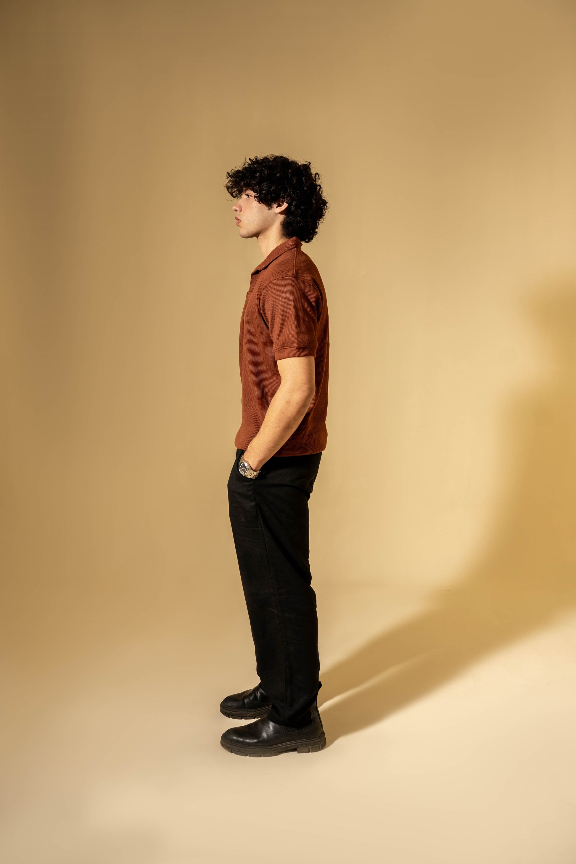 Man wearing a brown shirt and black pants standing against a beige wall.