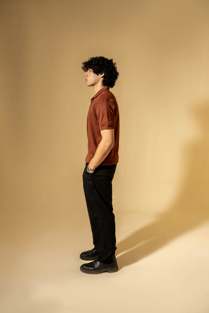 Man wearing a brown shirt and black pants standing against a beige wall.