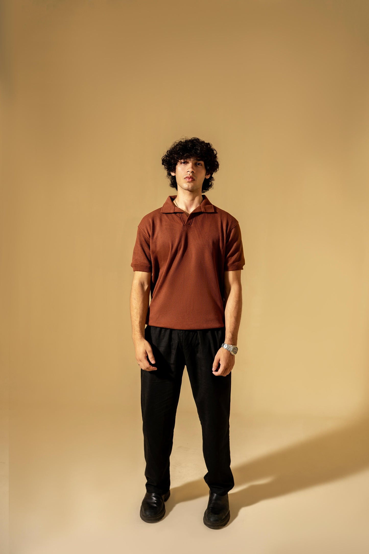 Man wearing a brown polo shirt and black pants against a beige background