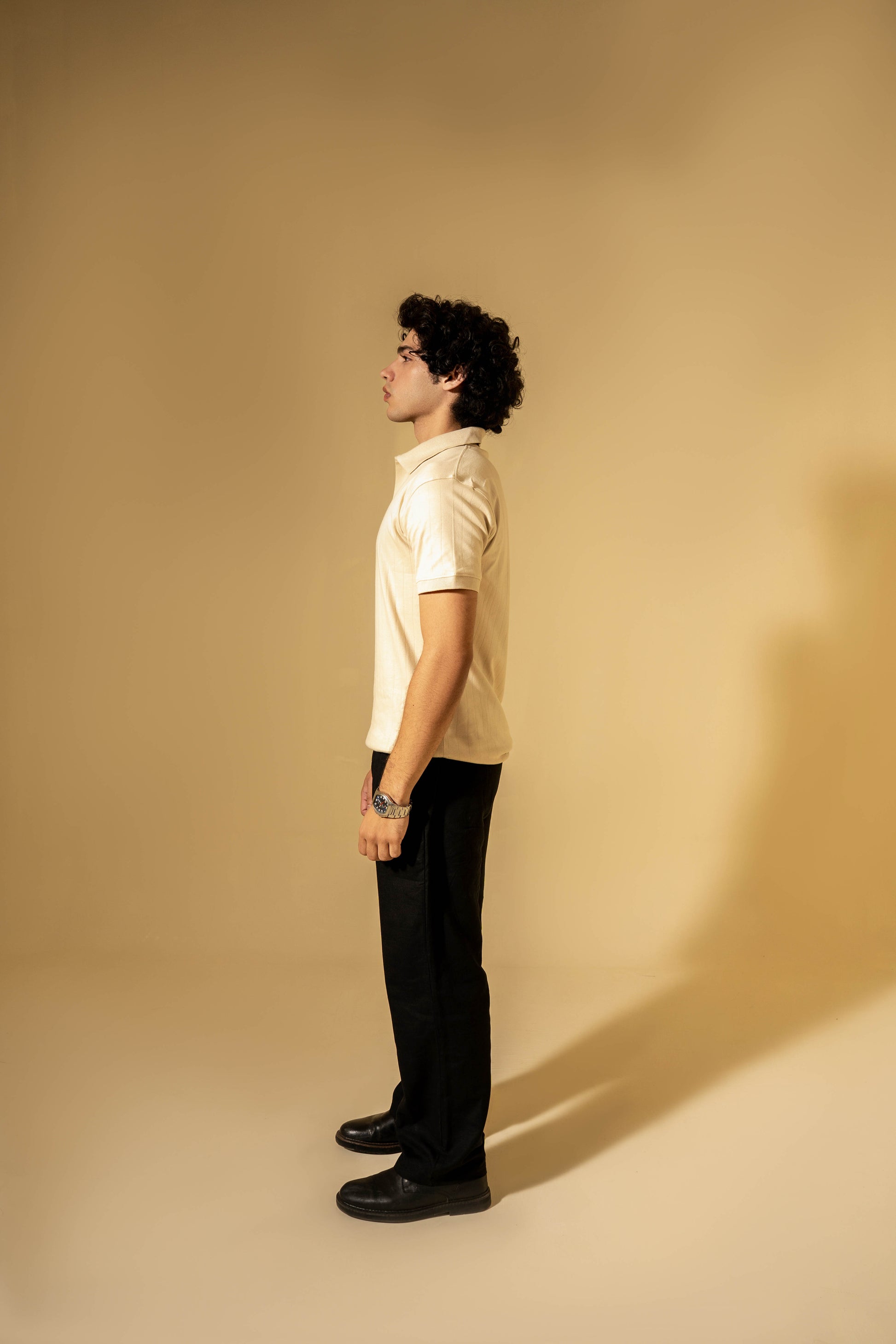 Person standing against a beige wall wearing a light-colored shirt and dark pants.